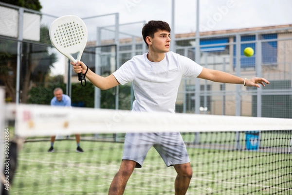 Fototapeta Portrait of emotional determined young guy playing padel tennis on open court in summer, swinging racket to return ball over net. Sportsman ready to hit volley