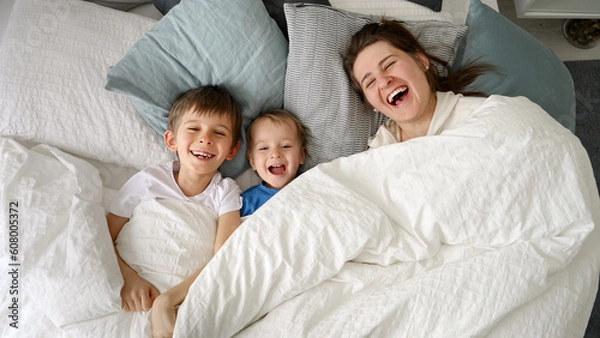 Fototapeta Happy laughing family with two kids hiding under blanket in bed and throwing it off. Concept of family happiness, relaxing at home, having fun in bed, parent and cheerful kids