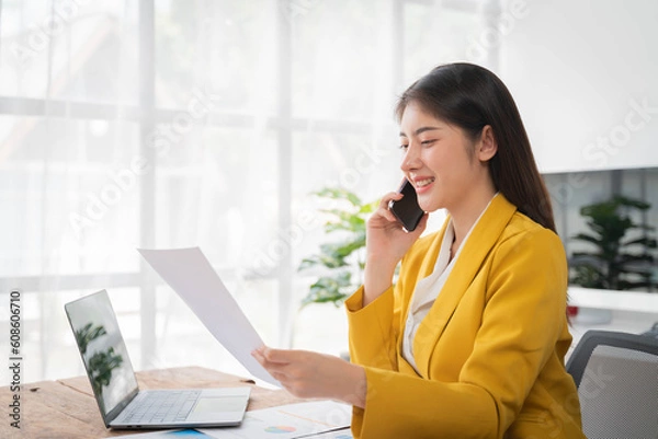 Obraz Businesswoman talking on cellphone holding paper sitting at desk and using laptop computer working online in office. Entrepreneurship and business career, distance freelance job concept.