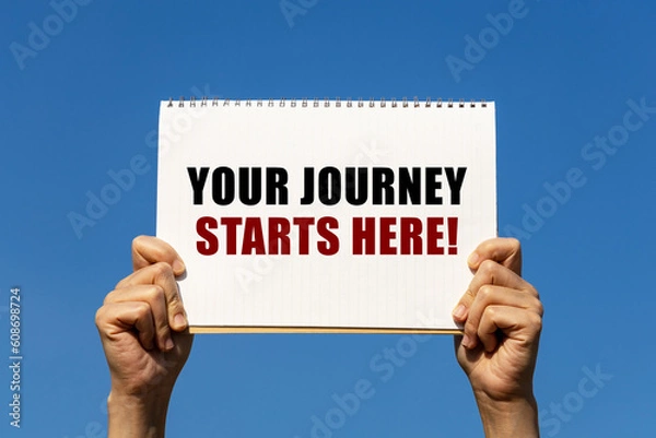 Fototapeta Your journey starts here text on notebook paper held by 2 hands with isolated blue sky background. This message can be used as business concept about your journey starts here.