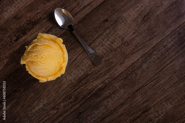 Fototapeta Tasty yellow cream ice cream served in a white bowl next to a spoon. Top photo with elements in the upper left corner of the image and empty space on the right. Ice cream ball.