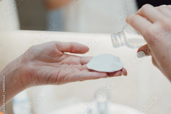 Obraz After showering the body and head wrapped in a towel, women's hands apply a moisturizing skin lotion to a cotton pad to wipe the face.