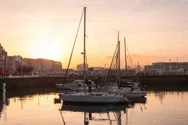 Fototapeta horizontal photo of the marina at sunset full of sailboards, yachts, boats and vessels with reflections on the water of sundown sky light and silhouettes of buildings at background