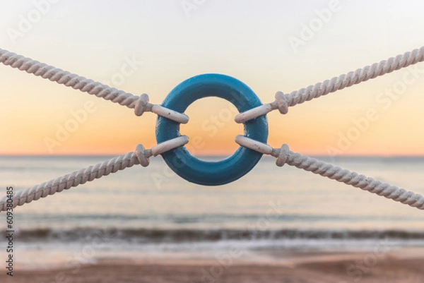 Fototapeta Fence of the Pedestrian way of Salinas, Asturias, with white sailboat ropes and a blue life ring, and at the background a blurry sand beach with quiet waves from the Atlantic Ocean at sundown