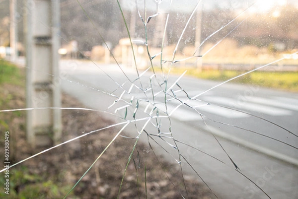 Fototapeta A glass window of a car punched by an angry people who hits a punch, a blurry background of a street with road and grass on a summer day