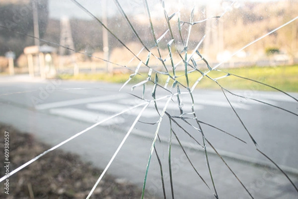Fototapeta A glass window of a car punched by an angry people who hits a punch, a blurry background of a street with road and grass on a summer day 