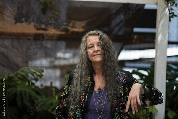 Fototapeta With one hand on a fence and a sun shade fabric in the background, a person relaxes in their backyard. They have flowing gray hair and a nose piercing.
