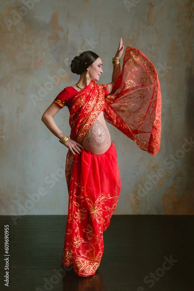 Obraz Pregnant woman in red sari. mehendi. Indian painting on woman's hands and pregnant belly with henna tattoo on room background