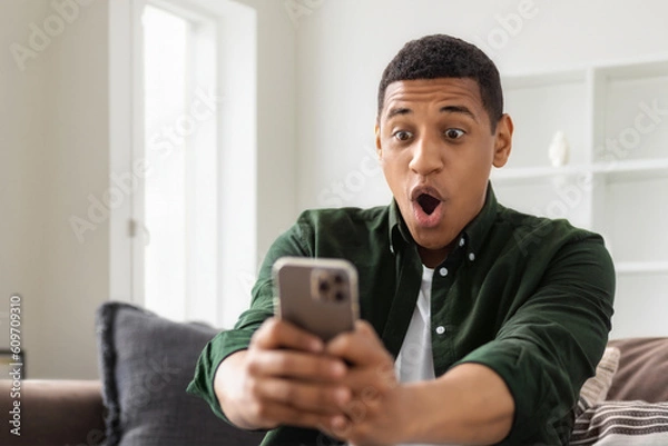 Fototapeta Happy excited African American man holding a mobile phone in his hands, looking at the screen with a surprised face, young male reading good news sitting at home on the couch