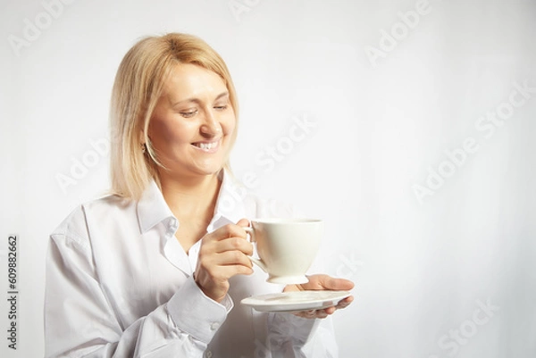Fototapeta Portrait of pretty blonde smiling woman posing with cup of tea or coffee on white background. Happy girl model in white shirt in studio. The concept of pleasant morning at home or at work. Copy space