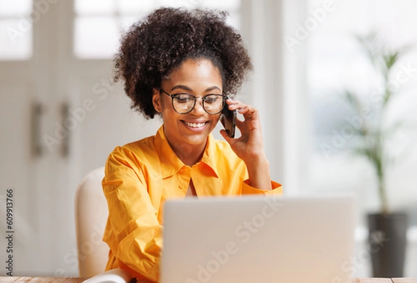 Fototapeta Beautiful young smiling ethnic woman making call via smartphone while working remotely from home while sitting at desk and talking to coworkers .