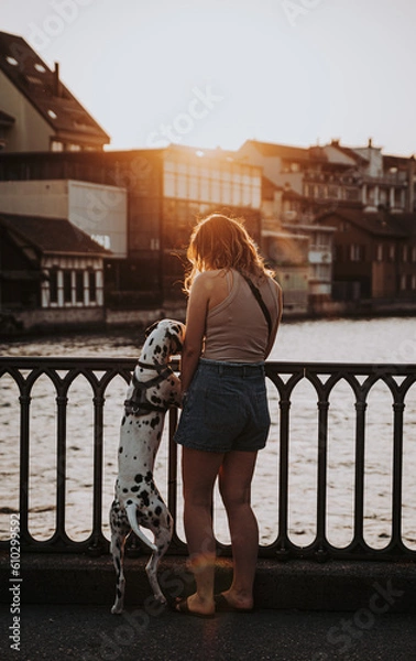 Fototapeta Frau steht mit Hund an Geländer bei Sonnenuntergang am Fluss
