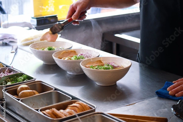 Fototapeta Bowls of ramen on a countertop in a kitchen with steam smoke of boiling noodles, hands put ingredients into a bowl with all supplies to preparing Japanese ramen to serve in a restaurant