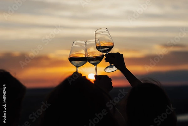 Fototapeta Close-up of hands cheers with glasses of wine at amazing sunset. Clinking glasses at sunset.