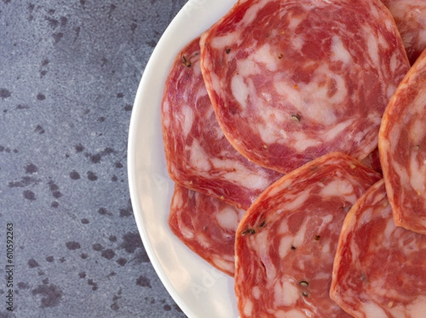 Obraz Top close view of several slices of sweet sopressata on a white plate atop a gray mottled background.