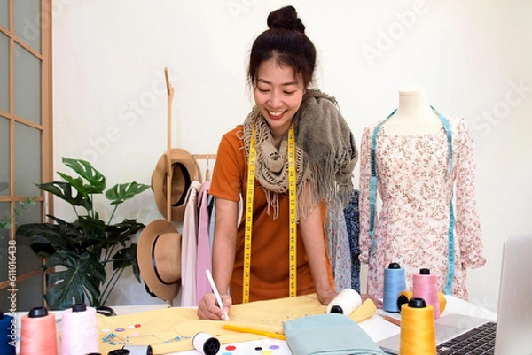 Fototapeta Asian woman designer for fashion working in the showroom and fashion working space. Lifestyle stylish use laptop for designing in studio, fashion business Concept