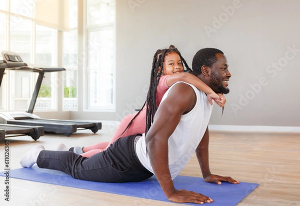 Fototapeta Father doing yoga with cute daughter, beginner doing simple exercise on purple background in gym.