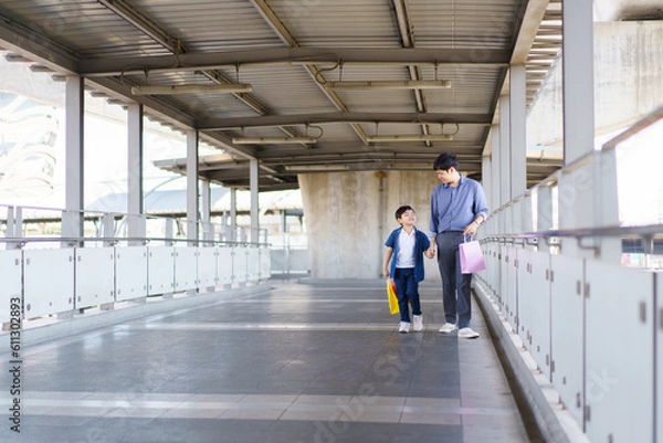Fototapeta Happy cheerful Asian little boy and his father walking in the city with carrying a colorful paper shopping bag, father and son walking together in the city or downtown.