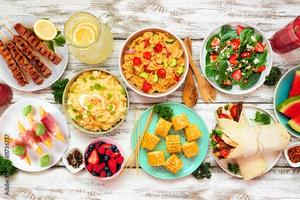 Fototapeta Summer food table scene over a white wood background. Collection of refreshing salads, fruit, wraps and BBQ grilled skewers. Top view.