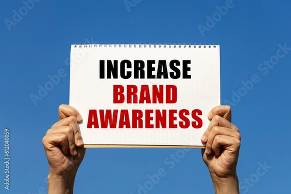 Fototapeta Increase brand awareness text on notebook paper held by 2 hands with isolated blue sky background. This message can be used as business concept about increase brand awareness.
