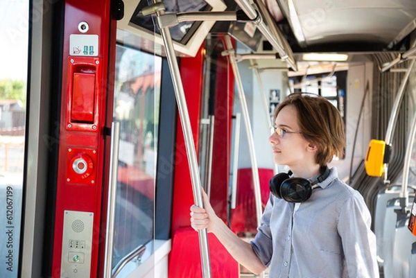 Obraz Tennager boy with headphones travels independently by public transport. Guy staying near an exit door in a tram, looking outside in a window. Way to the school. Eco-friendly transportation in a Europe