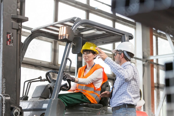 Fototapeta Supervisor directing worker driving forklift in factory