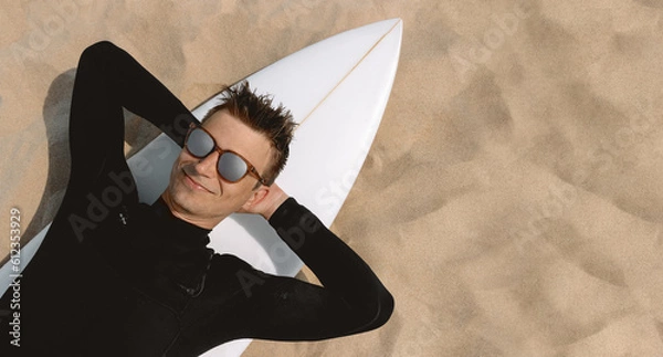 Fototapeta Surfer in wetsuit and sunglasses with surfboard relaxing at beach, sand background. View from above and copy space for text. 