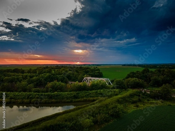 Fototapeta Aerial shot of a cloudy summer sunset over a railroad bridge and a green landscape