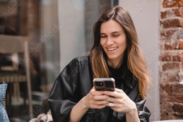 Fototapeta Portrait of happy female dressed casually holding mobile phone, typing messages, communicating with friends via social networks, using high Internet connection at cafe. Woman look flirting.