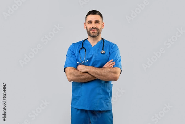 Fototapeta Middle aged man doctor in uniform with crossed arms on chest looking at camera on light gray background, free space