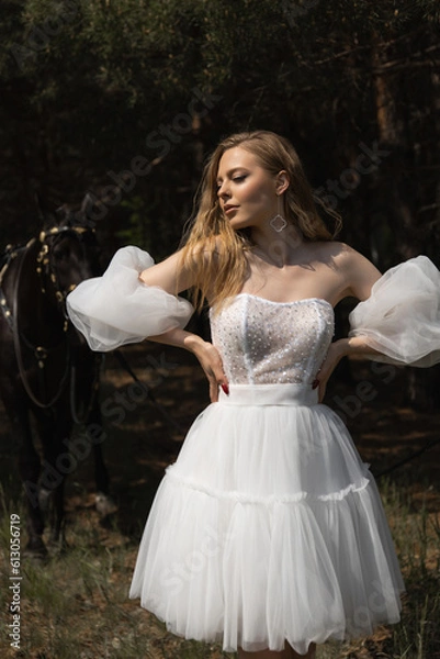 Obraz A beautiful Caucasian girl in a wedding dress stands in the forest next to a horse.