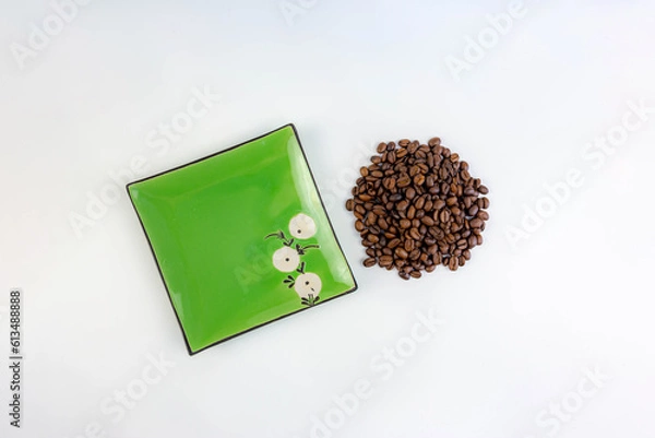 Fototapeta A pile of coffee beans next to a square green saucer, white background