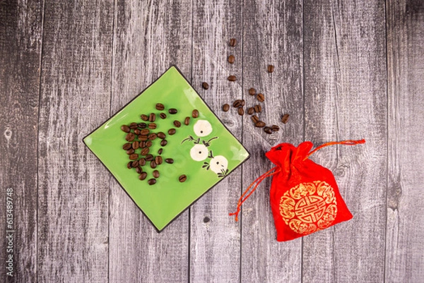 Fototapeta Coffee beans on a green saucer with a red bag next to it, wooden background, photo from above