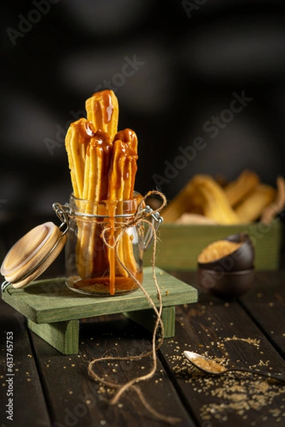 Fototapeta Churros served in a glass jar with caramel sauce, placed on a rustic wooden surface. Crispy golden pastry sticks, sugar crumbs, and warm tones create a cozy dessert atmosphere.