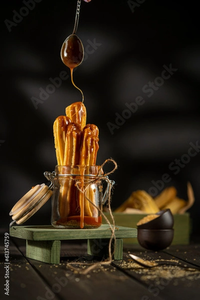 Fototapeta Churros served in a glass jar with caramel sauce, placed on a rustic wooden surface. Crispy golden pastry sticks, sugar crumbs, and warm tones create a cozy dessert atmosphere.