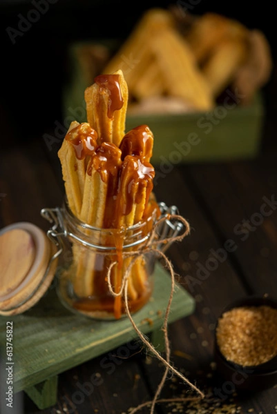 Fototapeta Churros served in a glass jar with caramel sauce, placed on a rustic wooden surface. Crispy golden pastry sticks, sugar crumbs, and warm tones create a cozy dessert atmosphere.