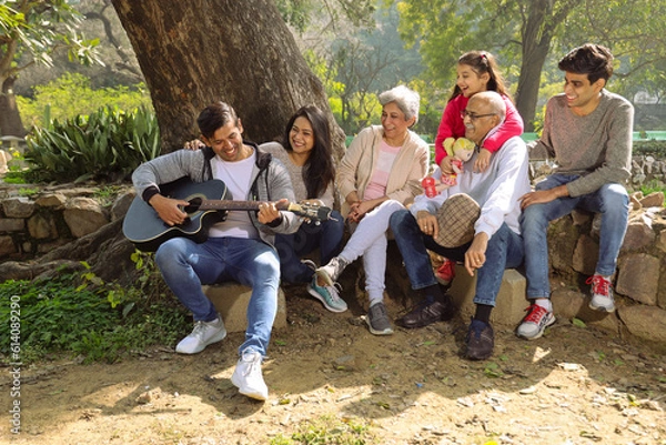 Obraz Happy Indian big family sitting on a pedestal under the shades of a tree and watching a family member playing guitar.