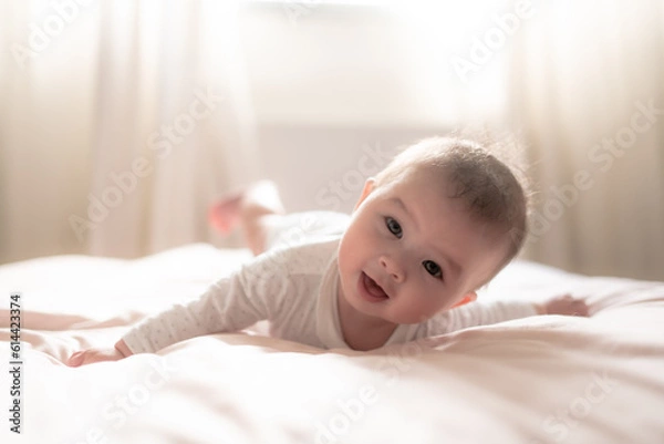 Fototapeta adorable and happy 5 months old baby girl gleefully discovers boundless joy while playfully exploring her bed. Surrounded by the warm glow of natural light the little on smiles cheerfully
