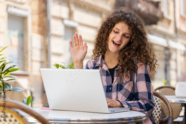 Fototapeta Portrait of pretty young woman using laptop having video call chat conversation for online meeting and presentation outdoors. Girl tourist recording blog sitting in urban sunshine city cafe terrace