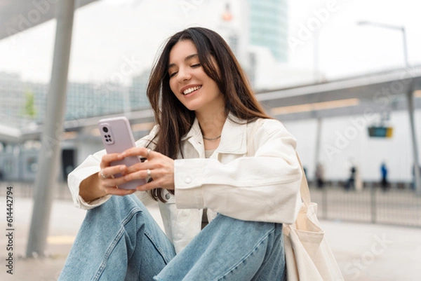 Fototapeta Bright and cheerful young woman with shopper on shoulder waiting public transport at station smiling beautifully while texting on phone