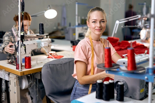 Fototapeta Dressmakers women sewing clothes on sewing machine in factory together, at work. Attractive ladies enjoy tailoring, making clothes, blonde caucasian craftswoman looking at camera smiling