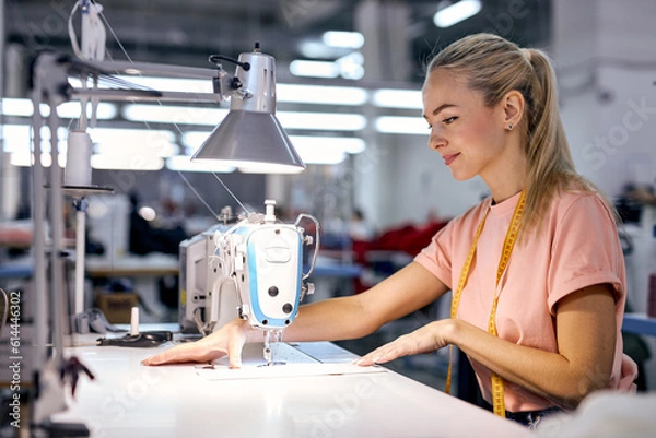 Obraz concentrated blonde caucasian woman seamstress sits using modern sewing machine. lady working in textile workshop. Tailoring, sewing as a small business or hobby concept. sewing process