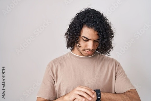 Fototapeta Hispanic man with curly hair standing over white background checking the time on wrist watch, relaxed and confident