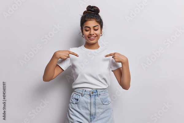 Fototapeta Young dark haired woman captures attention with her vibrant energy with contagious smile directs focus to her blank white tshirt using two fingers shows place for your logo or design poses indoor