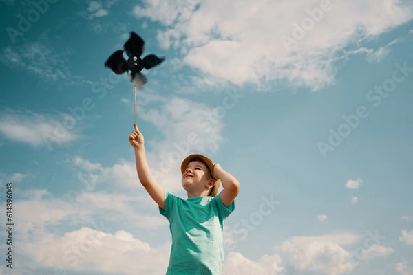 Fototapeta Little boy holds handcrafted windmill pinwheel in hand against the background of sky and clouds	