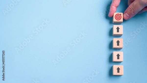 Fototapeta Hand holding wooden block with target aiming dart board and arrow up on blue background. Business achievement objective goal and project progress growth value processing marketing financial concept