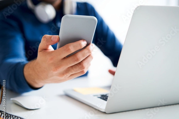 Fototapeta Closeup of man holding smartphone next to laptop screen sitting at a desk