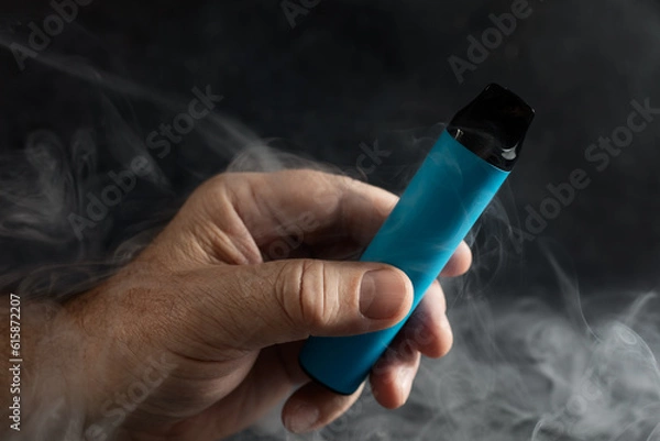 Fototapeta disposable electronic cigarette in a man's hand on a dark background with smoke around. The concept of modern alternative smoking, vaping and nicotine