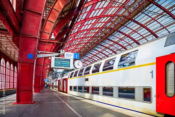 Fototapeta Antwerp, Belgium. Central indoor railway station. Platform made of red metal constructions with clock and panel with departure or arrival schedule. Modern double decker high-speed train