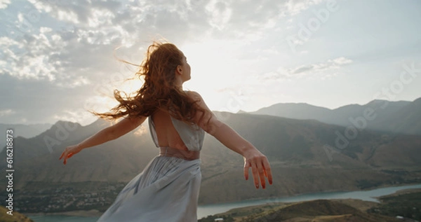 Obraz Woman in white dress standing on top of a mountain with raised hands while wind is blowing her dress and red hair - freedom, nature concept 